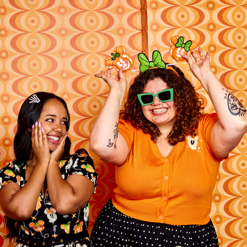 Two women hanging out against an orange background, one on the left wearing our Pumpkin Balloon Allison Dress and one on the right wearing the Pumpkin Balloon Domonique Cardigan with a Pumpkin Ear Headband 